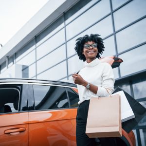 Young african american woman in glasses stands outdoors near modern car after shopping with packages